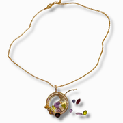 A gold chain necklace with a circular pendant featuring a clear dome and colorful inlaid stones, plus several loose multicolored gemstones displayed beside it on a white background.