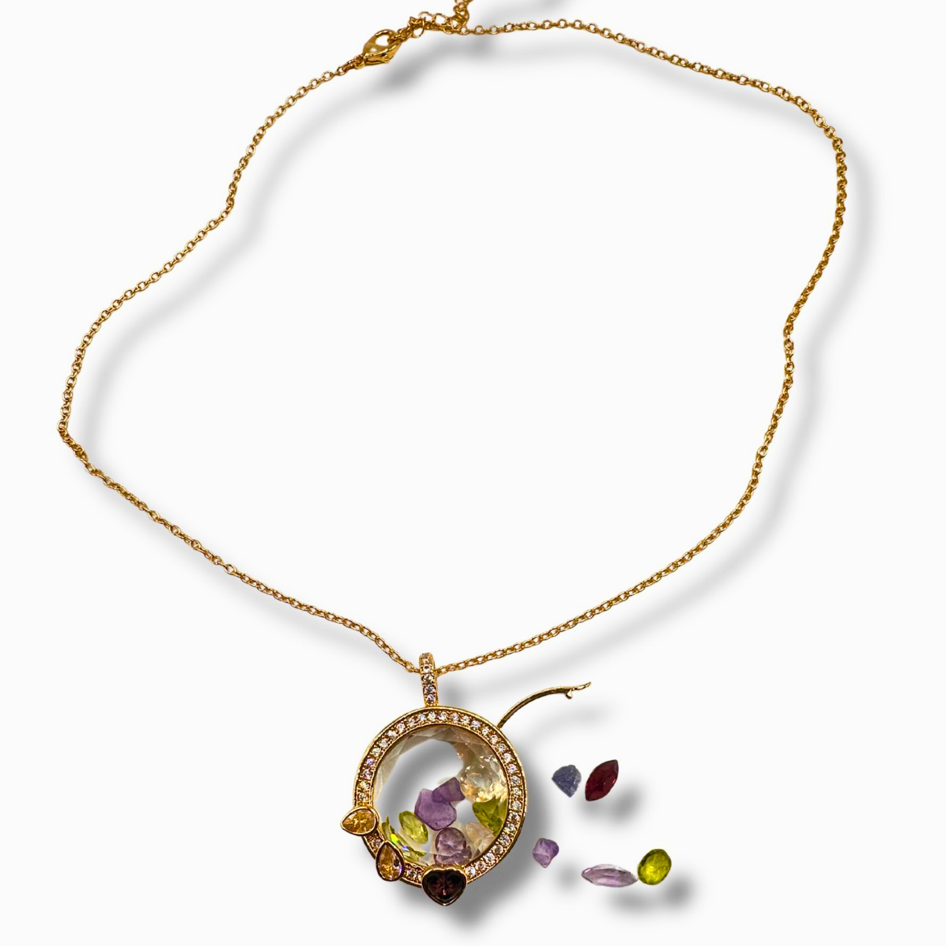 A gold chain necklace with a circular pendant featuring a clear dome and colorful inlaid stones, plus several loose multicolored gemstones displayed beside it on a white background.