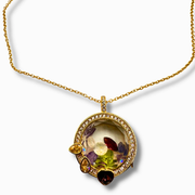 A gold pendant necklace featuring a round, openwork design filled with dried botanical elements, surrounded by small clear stones and accented with colorful gemstones in yellow, purple, and dark red, shown on a white background.
