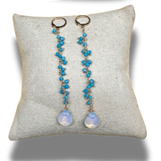 Pair of earrings with turquoise stones and opal-like beads on a textured gray pillow.