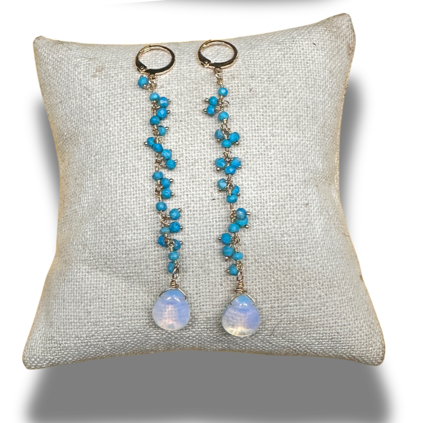 Pair of earrings with turquoise stones and opal-like beads on a textured gray pillow.